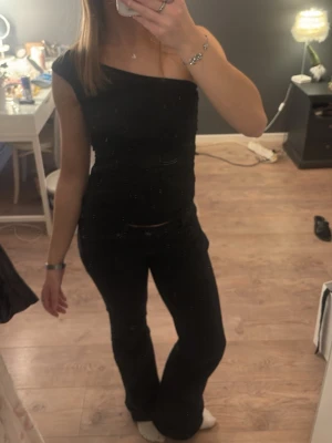 Svart oneshoulder topp - Säljer en svart oneshoulder topp med tight passform. Toppen är ärmlös på ena sidan och har en axelrem på andra sidan, vilket ger en asymmetrisk look. Materialet känns mjukt och stretchigt, perfekt för en trendig outfit.