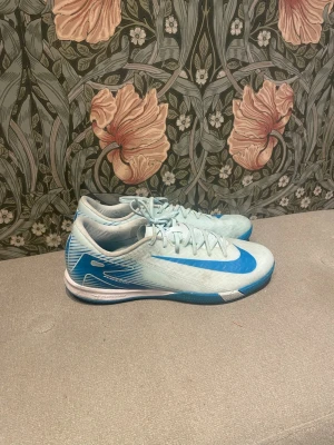 Nike blå fotbollsskor - Säljer ett par ljusblå Nike fotbollsskor med tydliga blå detaljer och snörning. Skorna har en låg profil och platt sula, perfekt för spel på inomhusplan. Tillverkade i syntetmaterial för lätt känsla och bra grepp. Hör gärna av dig vid frågor!