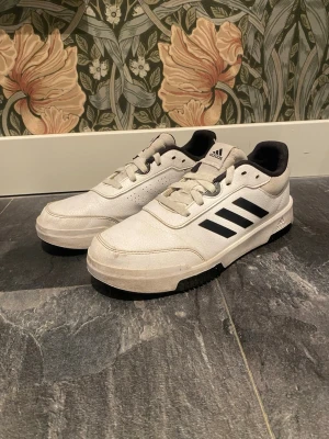 Vita Adidas sneakers med svarta ränder - Säljer ett par klassiska vita Adidas sneakers med svarta ränder på sidan och svart detalj på hälen. Skorna har snörning, platt sula och är tillverkade i syntetmaterial med textilfoder. Perfekta för dig som gillar stilrena och sportiga skor.