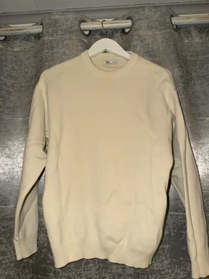 Beige stickad tröja från Zara - Säljer en enkel och stilren beige stickad tröja från Zara. Tröjan har rund halsringning, ribbade muddar vid ärmslut och nederkant samt lång ärm. Perfekt för lager på lager under höst och vinter.