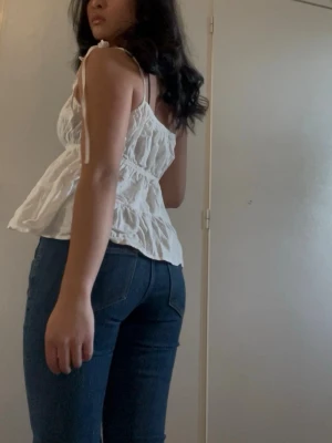 Vit volangig linnetopp med knytband - Supersöt vit linnetopp med volanger och knytband på axlarna. Toppen har en luftig och flowy passform, smockad midja och är ärmlös. Perfekt för varma dagar och ger en romantisk vibe.