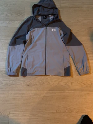 Grå vindjacka från Under Armour  - Snygg grå vindjacka från Under Armour med huva och dragkedja framtill. Jackan har mörkare grå detaljer på axlar, ärmar och rygg samt logga på bröstet och text på ryggen. Perfekt för utomhusaktiviteter och träning. Storlek M passar som L Pris kan diskuteras