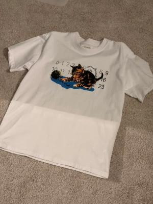 Vit t-shirt Maison Margiela kattmotiv - Vit t-shirt från Maison Margiela med tryck av en katt som leker med ett garnnystan och siffror i bakgrunden. Klassisk passform med rund hals och korta ärmar. Materialet är mjuk bomull och trycket har inslag av blått, svart och brunt.