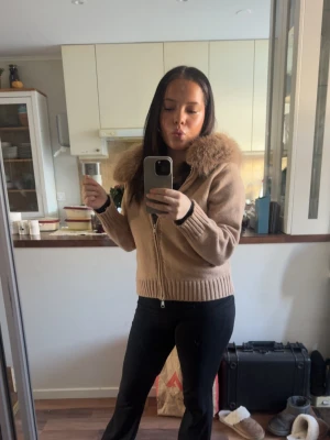 Beige stickad jacka med fuskpäls - Säljer min beiga jacka med fuskpäls i huvan, mysig runt våren och bra att styla med till vad som helst.