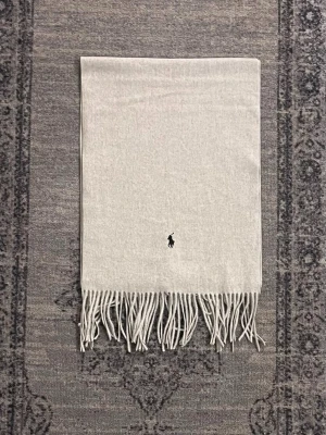 Grå och svart halsduk Polo Ralph Lauren - Två klassiska halsdukar från Polo Ralph Lauren. En i ljusgrått med broderad svart logga och en i svart med röd logga. Båda har långa fransar i ändarna och är tillverkade i mjuk ull. Perfekt accessoar för kyliga dagar.pris går att diskutera 