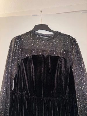 Svart glittrig långklänning i sammet - Supersnygg svart långklänning med glittriga prickar på ärmar och överdel. Klänningen har långärmade transparenta ärmar med volang vid ärmslut och en mjuk sammetskjol. Perfekt för dig som vill sticka ut med en unik look!   Står XL på klänningen men passar för L