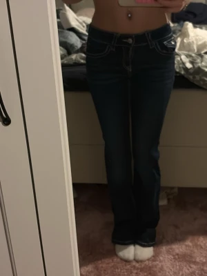 Mörkblå bootcut jeans med kontrastsömmar - Säljer ett par mörkblå jeans med tydliga vita kontrastsömmar och snygga fickdetaljer bak. Modellen har bootcut-ben och låg midja, vilket ger en klassisk och trendig siluett. Materialet är jeans och passformen är normal. Perfekta till sneakers eller boots.