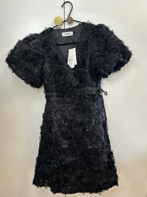 Svart blommig kortklänning Kappahl - Svart kortklänning från Kappahl med 3D-blommor. Klänningen har puffiga korta ärmar, omlott-v-ringning och knyt i sidan. Perfekt för dig som vill sticka ut med en unik och trendig look.