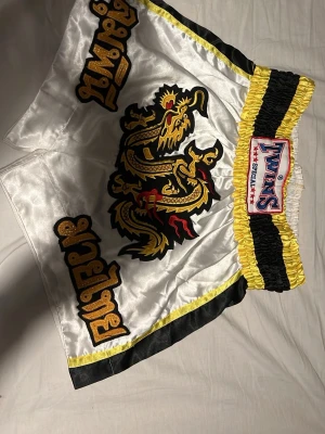 Twins Special Muay Thai shorts XL - Snygga Muay Thai shorts från Twins Special i blankt vitt tyg med svarta och gula detaljer. Shortsen har en stor broderad drake framtill och thailändsk text i guld. Bred resår i midjan för extra komfort och klassisk thaiboxningsstil.