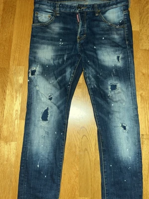 Blå Dsquared2 ICON jeans med slitningar - Säljer ett par blå Dsquared2 ICON jeans med slitna detaljer, hål och färgstänk för en riktigt edgy look. Jeansen har klassisk femficksmodell, normal passform och tydliga slitningar framtill och baktill. Tillverkade i jeansmaterial med ikonisk patch bak i midjan.