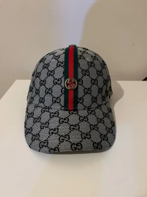 Grå Gucci keps med GG-mönster - Snygg keps från Gucci i grått med klassiskt GG-mönster över hela kepsen. Framtill går ett grönt och rött band med metallogo. Justerbar rem baktill med metallspänne där det står Gucci. Perfekt accessoar för att lyfta din outfit.