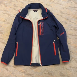 8848 Altiitude Blå vindjacka/skidjacka  med röda detaljer  - Snygg marinblå vindjacka från 8848 Altitude med röda dragkedjor och detaljer. Jackan har hög krage, huva och flera fickor med dragkedja. Insidan är ljusbeige och materialet känns slitstarkt och sportigt. Perfekt för dig som gillar en aktiv stil.