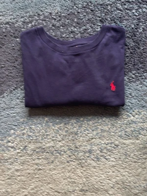 Mörkblå t-shirt från Polo Ralph Lauren - Snygg mörkblå t-shirt från Polo Ralph Lauren med klassisk röd logga broderad på bröstet. Tillverkad i mjuk bomull och har en normal passform med rund hals. Perfekt till jeans eller shorts för en clean och enkel stil. Nypris: 600kr 