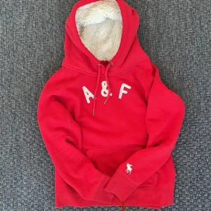 Fodrad Abercrombie & Fitch vintage hoodie - Snygg röd hoodie från Abercrombie & Fitch med fluffigt foder i huvan och broderad vit älg på ärmen. Stor A&F-logga framtill, snörning vid huvan och klassisk känguruficka. Perfekt till höst och vinter🤩. Storlek L på etiketten men passar xs-s  Säljs ej längre i butik och är därav sällsynt .Skriv vid funderingar 📲
