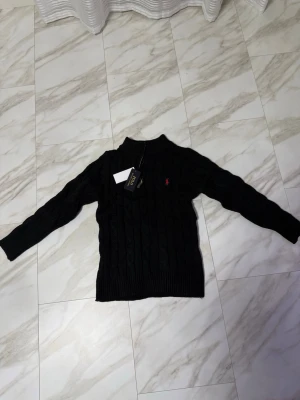 Svart stickad tröja Polo Ralph Lauren - Kabelstickad svart tröja från Polo Ralph Lauren med halv dragkedja i halsen och röd broderad logga på bröstet. Tröjan har ribbade muddar och lång ärm, perfekt för kyliga dagar. Klassisk och stilren design som funkar till allt.