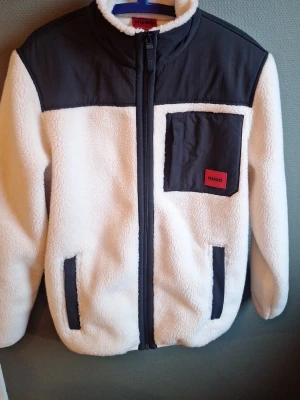 Vit teddyjacka från HUGO - Säljer en jätte fin teddyjacka från HUGO i vit och svart. Jackan har svart axelparti, bröstficka med röd HUGO-logga och dragkedja framtill. Fodrad med mjuk fleece och har två sidofickor. Perfekt för kalla dagar och ger en clean streetstyle. Tog 36 på storleken, står Storlek S i jackan.