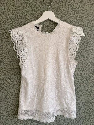 Vit spetsblus med volangärm - Supersöt vit blus i spets med blommigt mönster och volangdetaljer vid ärmarna. Blusen har hög hals med liten kant och är ärmlös med breda spetsvolanger. Perfekt för sommarens alla tillfällen och ger en romantisk vibe.Men tyvär ett litet hål 
