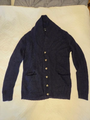 Mörkblå kofta från Filippa K, M - Mysig mörkblå kofta från Filippa K i storlek M. Koftan är stickad i en ullblandning med ribbade muddar, bred krage och knappar framtill. Två fickor framtill ger en chill vibe och den passar perfekt till lager på lager under höst och vinter.