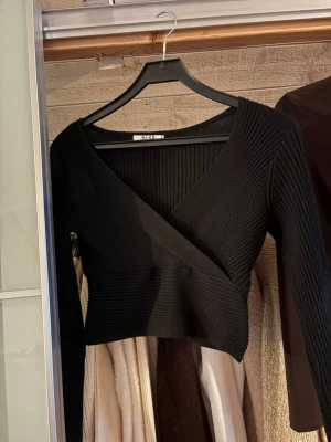 Svart omlottop från NA-KD - Säljer en svart ribbad omlottop från NA-KD. Tröjan har lång ärm, djup v-ringning och croppad passform. Perfekt till höst och vinter för en stilren look. Materialet är stretchigt och mjukt, vilket gör den riktigt bekväm.