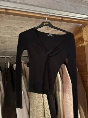 Svart omlottopp från Bershka - Säljer en svart omlottopp från Bershka med lång ärm och v-ringad hals. Toppen har en twistad detalj framtill som ger en snygg look. Materialet känns mjukt och stretchigt, perfekt att styla med jeans eller kjol.