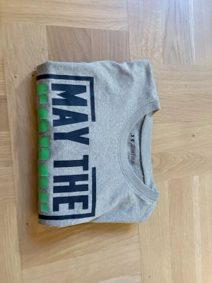 Grå Star Wars t-shirt Under Armour träning  - Säljer en grå t-shirt från Under Armour i samarbete med Star Wars. Framsidan har trycket 'May the Force be with you' i svart och neon-grönt. Klassisk rund hals och korta ärmar. Perfekt för dig som gillar coola prints och Star Wars-vibbar.