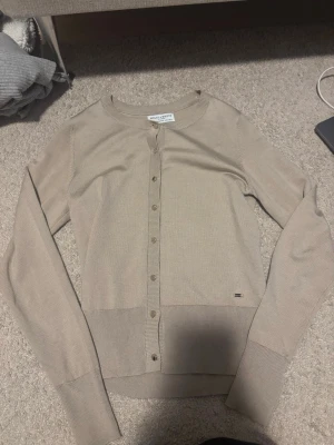 Beige kofta från Holly & Whyte - Säljer en stilren beige kofta från Holly & Whyte. Koftan har rund halsringning, långa ärmar och knappar framtill. Tillverkad i mjukt material som passar perfekt till lager på lager. Klassisk och enkel design med liten metallogo vid nederkanten.