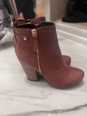 Bruna stöv ink stövletter från tiamo  - Snygga bruna stövletter från Tiamo med chunky klack och guldfärgad dragkedja på sidan. Skorna är gjorda i syntetmaterial med rund tå och har en stilren design som passar till många outfits. Perfekta för dig som vill ha både höjd och komfort.