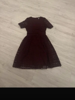 Vinröd spetsklänning med kort ärm - Vinröd klänning i spets med korta ärmar och rund hals. Kjolen är utställd och klänningen har ett vackert spetsmönster över hela plagget. Midjan är markerad och tyget är lätt transparent i detaljerna. Perfekt för dig som vill ha en elegant look.