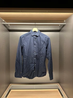 Mörkblå prickig skjorta Riley - Snygg mörkblå skjorta från Riley i slim fit med små vita prickar över hela tyget. Klassisk krage, långa ärmar och vita knappar framtill. Perfekt för dig som gillar stilrena detaljer och en smalare passform.