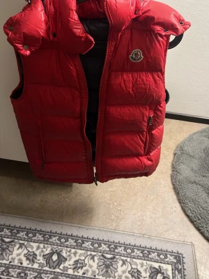 Röd dunväst från Moncler - Säljer en röd dunväst från Moncler med ikonisk logga på bröstet och coolt serietryck på insidan. Västen har hög krage, dragkedja framtill och två fickor med dragkedja. Perfekt för lager på lager och riktigt snygg till streetwear. Skick 6/10 Storlek 2. Hmu för fler funderingar 