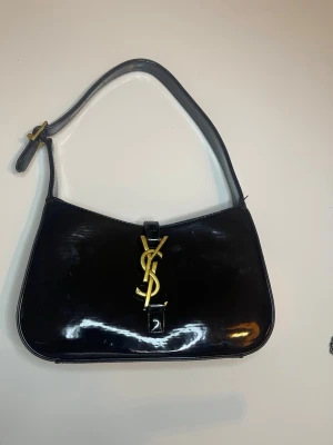 Svart axelväska från Yves Saint Laurent - Säljer en svart axelväska från Yves Saint Laurent med glansig finish och ikoniskt YSL-emblem i guld på framsidan. Väskan har justerbart axelband och är tillverkad i skinn. Perfekt storlek för det viktigaste och ger en lyxig vibe till din outfit.