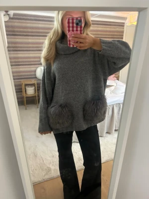Grå oversized stickad tröja med fuskpälsfickor - Mysig grå oversized stickad tröja med stor polokrage och två stora fickor i grå fuskpäls framtill. Perfekt för kalla dagar och ger en chill vibe. Långärmad och riktigt skön att bära till jeans eller leggings.