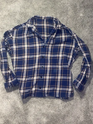 Flanelskjorta - Säljer en blå rutig skjorta med långa ärmar och knappar framtill. Skjortan har klassisk krage och är tillverkad i mjukt bomullstyg. Perfekt för dig som gillar avslappnad stil med en tidlös touch.