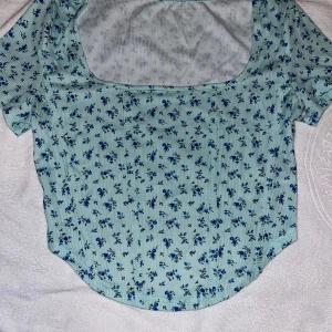 Mintgrön topp med kort ärm - Superfin topp i mintgrönt med små blå blommor och diskreta ränder. Modellen har korta ärmar, rundad nederkant och fyrkantig urringning framtill. Perfekt för dig som gillar en söt och fräsch stil med blommigt mönster. Aldrig använd