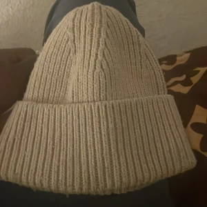 Beige stickad mössa från Object - Säljer en beige stickad mössa från Object. Mössan har en klassisk ribbstickad design med uppvikt kant och är tillverkad i mjukt material. Perfekt accessoar för kyliga dagar.