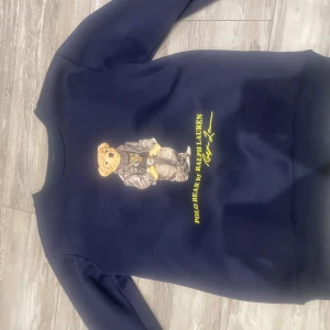Mörkblå Polo Bear tröja Ralph Lauren - Mörkblå sweatshirt från Polo Ralph Lauren med ikoniska Polo Bear-trycket framtill. Tröjan har rund halsringning, långa ärmar och mjukt material som känns skönt mot huden. Perfekt för dig som gillar klassisk och cool streetstyle.