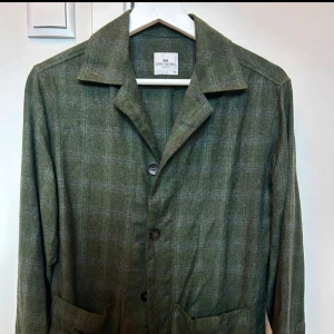 Grön rutig overshirt från John Henric - Stilren grön overshirt från John Henric i rutig design. Jackan har klassisk krage, knäppning framtill med fyra knappar och två stora fickor. Tillverkad i mjukt ullmaterial som ger en skön känsla och passar perfekt för lager-på-lager. Strl XS passar även S