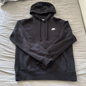 Svart Nike hoodie med huva och logga - Svart hoodie från Nike med klassisk vit logga på bröstet. Tröjan har huva med snörning, känguruficka och långa ärmar. Mjuk insida och ribbade muddar för extra komfort. Perfekt för chill eller streetwear.