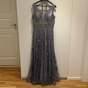 Glittrig långklänning i silvergrått - Säljer en magisk långklänning i silvergrått med glittriga detaljer och v-ringning. Klänningen har breda axelband, markerad midja med två rader av pärl- och paljettdekor samt ett skirt, glittrigt tyg med blommönster. Perfekt för bal eller fest!