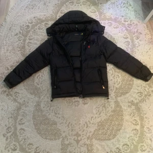 Svart pufferjacka från Polo Ralph Lauren - Svart pufferjacka från Polo Ralph Lauren med huva och dragkedja framtill. Jackan har två fickor med dragkedja och röd broderad logga på bröstet. Perfekt för kalla dagar och riktigt snygg till streetwear-stilen.