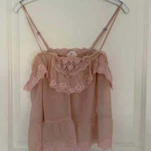 Rosa volangblus från Stradivarius - Supersöt ljusrosa blus från Stradivarius med tunna axelband, volanger och spetsdetaljer. Blusen är luftig och har broderade blommor längs kanterna samt en romantisk, flowy passform. Perfekt för sommarens varma dagar och ger en feminin vibe. Aldrig använd men saknar lapp.