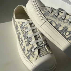 Christian Dior gråvita sneakers - Snygga sneakers från Christian Dior med grått och vitt monogrammönster, platt sula och rund tå. Skorna har snörning med vita skosnören och detaljer som 'Christian Dior' på snörningen. Tillverkade i textil med gummisula och cool stjärndetalj under sulan.