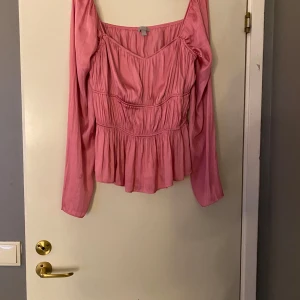 Rosa omlottblus med volang - Säljer en söt rosa blus med lång ärm och omlottvolang nedtill. Blusen har v-ringad hals och är gjord i ett mjukt, glansigt material som ger en lyxig känsla. Den har rynkade detaljer över hela framsidan och en flowy passform som ger en romantisk vibe.