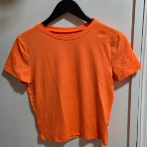 Orange croppad t-shirt, M - En enkel och fräsch orange croppad t-shirt med rund hals och korta ärmar. Toppen är i mjuk bomull och har en clean look utan tryck eller mönster. Perfekt att styla med jeans eller kjol för en chill vibe.