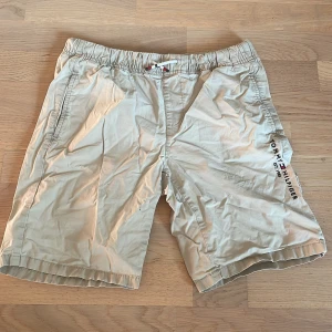 Beige shorts från Tommy Hilfiger - Snygga beige shorts från Tommy Hilfiger med elastisk midja och snörning. De har sidofickor, en bakficka och logotyptryck på benet. Perfekta för sommaren och har en clean, klassisk look som funkar till många stilar.