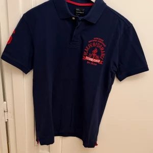 Marinblå pikétröja Peak Performance M - Marinblå pikétröja från Peak Performance i 100% bomull. Klassisk krage, kort ärm och röd brodyr på bröstet samt siffran 86 på ärmen. Snygga röda detaljer på insidan av kragen. Perfekt för en sportig och stilren look. Storlek M.