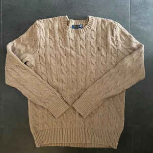 Beige kabelstickad tröja Polo Ralph Lauren - Kabelstickad beige tröja från Polo Ralph Lauren med klassisk rund halsringning och broderad logga på bröstet. Tröjan är stickad i bomull och har ribbade muddar vid ärmslut och nederkant. Perfekt för lager-på-lager under höst och vinter. Pris går att diskutera vid snabb affär! Och självklart är den äkta. 