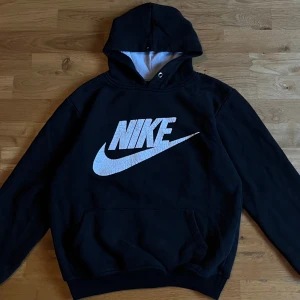 Svart Nike hoodie med stor logga - Svart hoodie från Nike med stor vit logga framtill. Klassisk känguruficka och huva med ljus insida. Mjuk och skön bomullsblandning, perfekt för chill eller träning. Långärmad och normal passform.