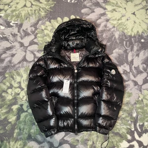 Svart pufferjacka från Moncler - Säljer en svart, glansig pufferjacka från Moncler med huva och dragkedja. Jackan har klassisk Moncler-logga på ärmen och coolt serietryck på insidan. Fylld med dun och har två fickor framtill. Perfekt för kalla dagar när du vill hålla stilen. Storlek 2/M Skick för mer bilder Nfc scan funkar på flera ställen‼️