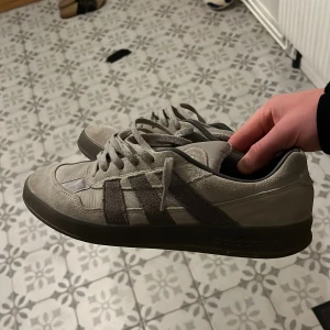 Adidas grå sneakers i mocka - Säljer ett par stilrena Adidas alhoa sneakers i grå mocka med klassiska ränder på sidan och broderad detalj på hälen. Skorna har snörning, platt sula och rund tå. Perfekta för dig som gillar en clean och enkel look med snygga detaljer.